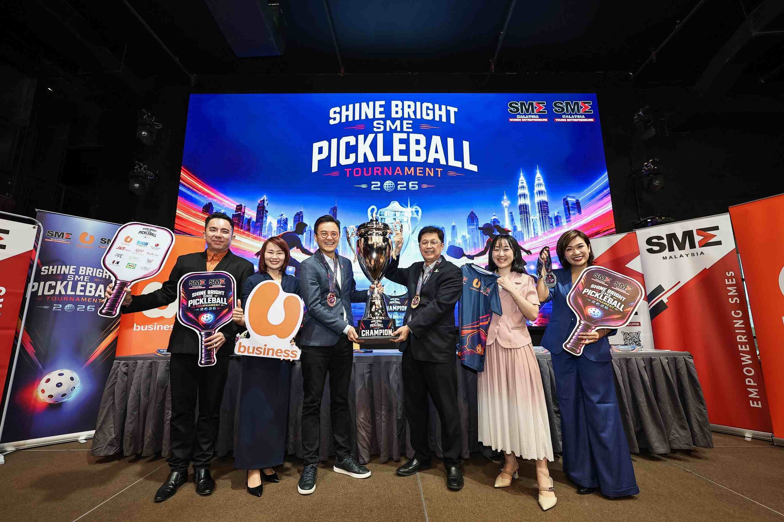SME Malaysia leaders and U Business partners posing on stage holding the championship trophy and branded pickleball paddle props