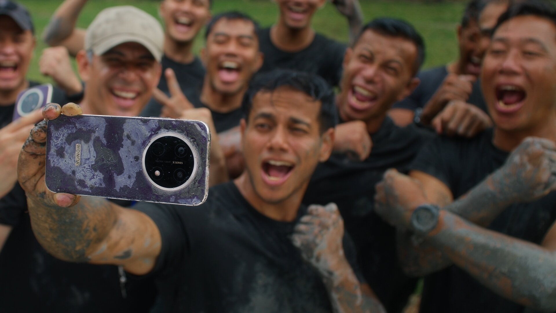 Group of mud-covered participants cheering while taking a selfie with a muddy HONOR X9e Pro smartphone