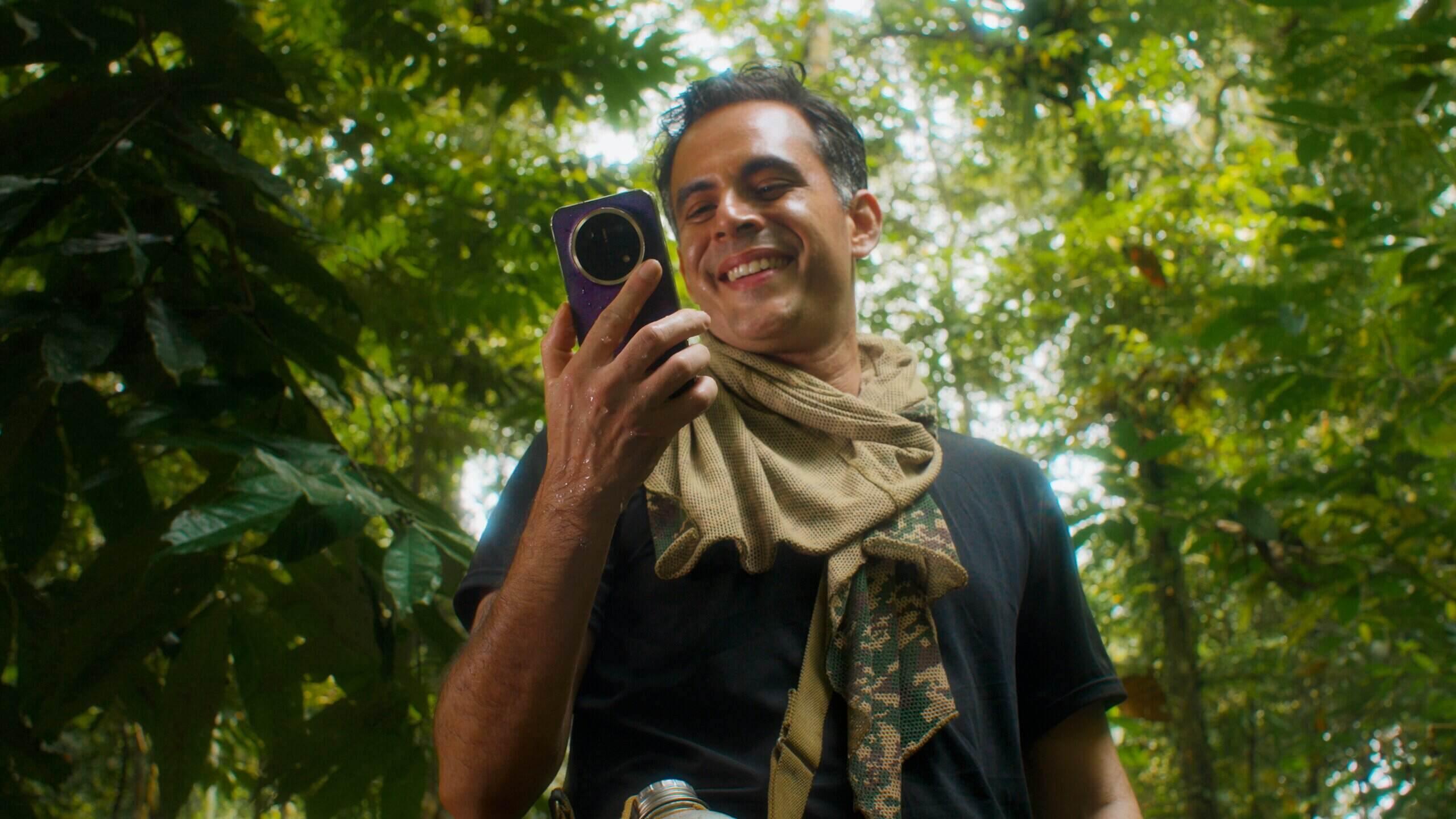 Man smiling and looking at a purple HONOR X9e Pro smartphone while standing in a tropical forest setting